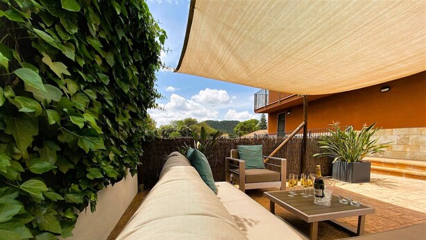 A covered outdoor seating area with a sofa, chairs, and a table, surrounded by greenery and a view of a building. - Morera Blanca (Canet de Fals)