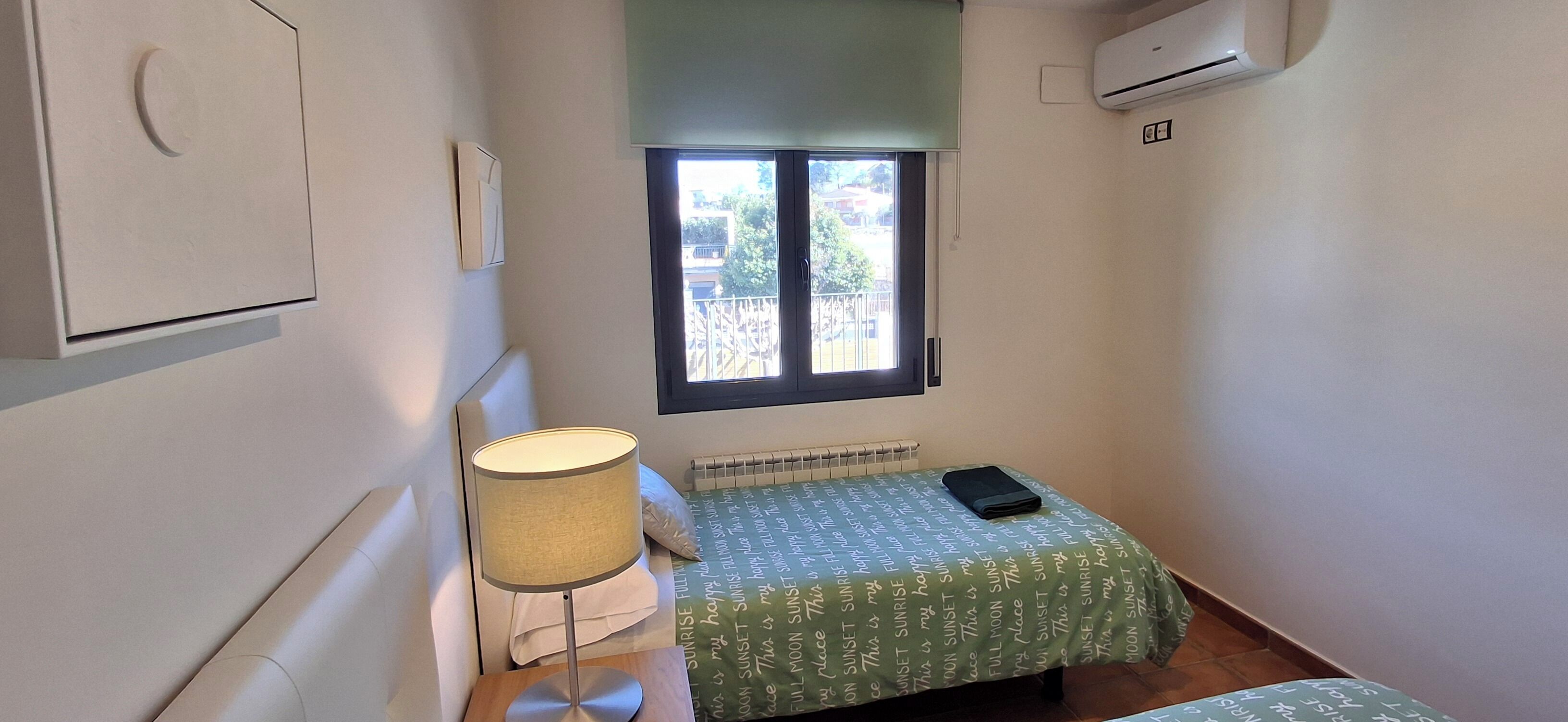 A small room with a bed, a lamp, a window, and an air conditioning unit.