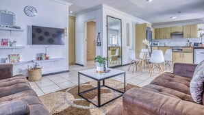 A modern living room with a sofa, a coffee table, and a TV. There is a dining area with chairs and a table. The kitchen is visible with cabinets and appliances. - The Cottage-1 Bed-pool-bbq-fibre-solar-samrand (Centurion)