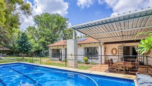 A swimming pool with a covered patio area, a dining table, and a view of a house with a tiled roof. - The Cottage-1 Bed-pool-bbq-fibre-solar-samrand (Centurion)
