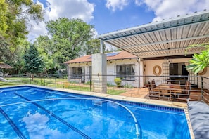 A swimming pool with a covered patio area, a dining table, and a view of a house with a tiled roof. - The Cottage-1 Bed-pool-bbq-fibre-solar-samrand (Centurion)