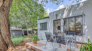 A patio with a table and chairs, a large tree, and a white building with a window and a door. - The Cottage-1 Bed-pool-bbq-fibre-solar-samrand (Centurion)