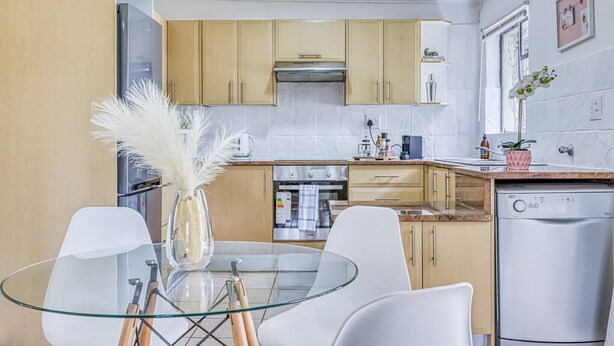A modern kitchen with wooden cabinets, a glass dining table, and a dishwasher. - The Cottage-1 Bed-pool-bbq-fibre-solar-samrand (Centurion)