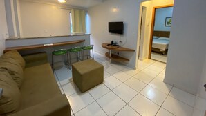 A modern living room with a sofa, a coffee table, and a TV mounted on the wall. There are three green chairs and a small table with a phone and charger. A door leads to a bedroom with a bed and a window. - Golden Dolphin 01 Quarto - Caldas Novas -GO (Caldas Novas)