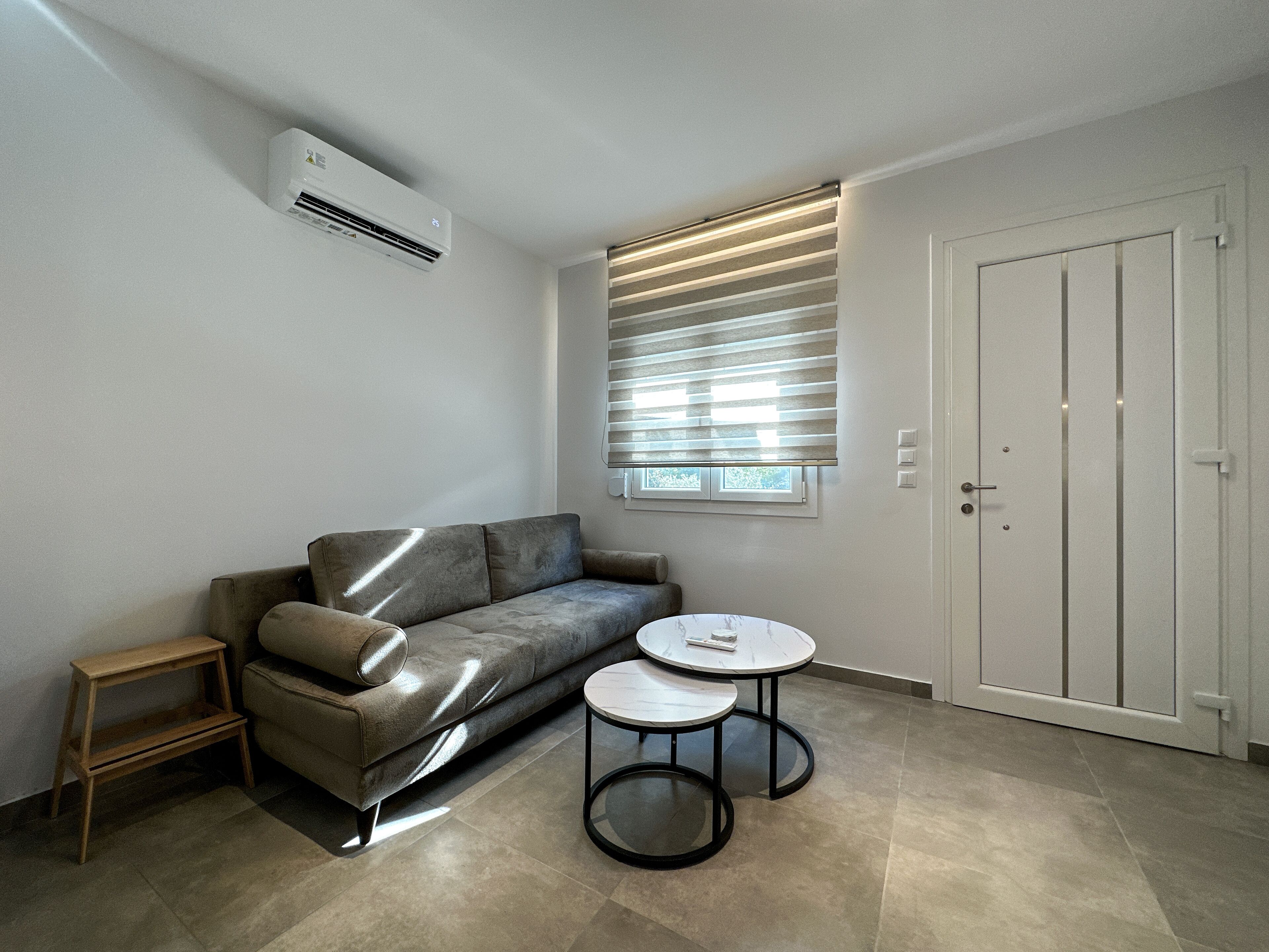 Uma sala de estar moderna com um sofá cinzento, uma pequena mesa de apoio de madeira, duas mesinhas redondas, uma porta branca e uma janela com persianas.