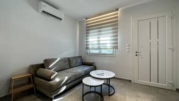 Uma sala de estar moderna com um sofá cinzento, uma pequena mesa de apoio de madeira, duas mesinhas redondas, uma porta branca e uma janela com persianas.