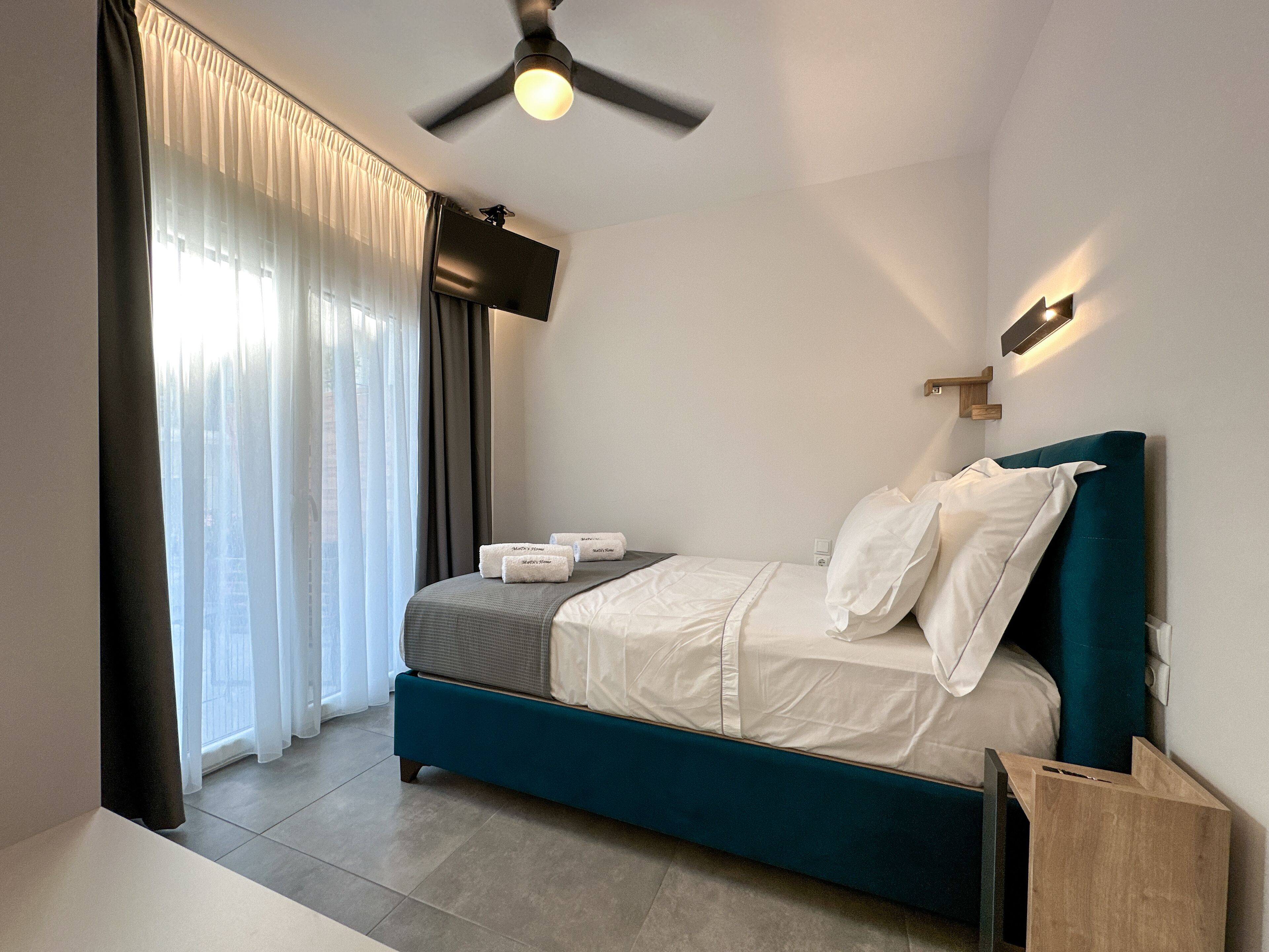 Ein modernes Schlafzimmer mit einem türkisfarbenen Bett, weißer Bettwäsche, einem flachbildfernseher an der Wand und einem Deckenventilator.