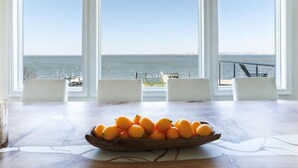 Una mesa de comedor con un cuenco de madera lleno de naranjas, tres sillas blancas y vistas al mar a través de amplias ventanas.