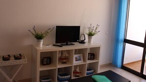 A room with a TV on a white stand, two potted lavender plants, a small table with a green mat, and a window with blue curtains. - Albufeira 2 - Beach & Old Town (Albufeira)