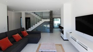A modern living room with a dark sofa, a flat-screen TV, and a staircase leading to another level. - Villa Goncinha - Private Pool & BBQ (Loulé)
