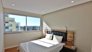 A bedroom with a large window, a bed with a wooden headboard, a bedside table, and a nightstand.