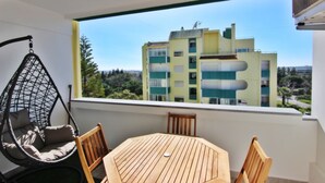 A balcony with a hanging chair, a wooden table, and a view of a multi-story building.