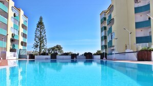 A swimming pool surrounded by multi-story buildings with balconies, a tall evergreen tree, and a clear blue sky.