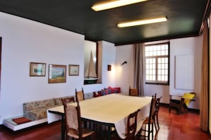 Un comedor con una mesa preparada para seis personas, rodeada de sillas de madera. Hay un sofá con cojines, una ventana con cortinas y obras de arte enmarcadas en la pared.