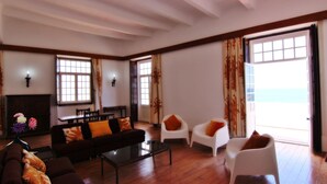 A spacious living room with a wooden floor, a dark sofa, a white armchair, and a coffee table. There is a fireplace with a vase of flowers, and large windows with curtains. - Casa dos Tios - Sea View, Beach & Old Town Center (Albufeira)