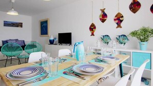 Comedor con mesa de madera dispuesta para cuatro personas, sillas blancas y un televisor sobre un mueble blanco.