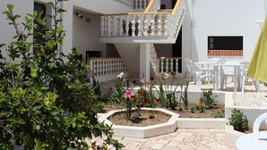 Un edificio de varios niveles con paredes blancas y balcones, zona de estar al aire libre con sillas de plástico blanco y un jardín con diversas plantas y flores.