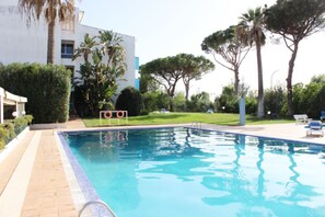 Una piscina con agua cristalina azul, rodeada por un edificio blanco, palmeras y un área verde.