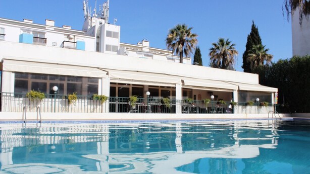 A swimming pool with a clear blue water surface, surrounded by a white building with multiple windows and balconies.