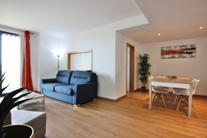 Een woonkamer met een bank, een eettafel met stoelen, een potplant en een schilderij aan de muur.