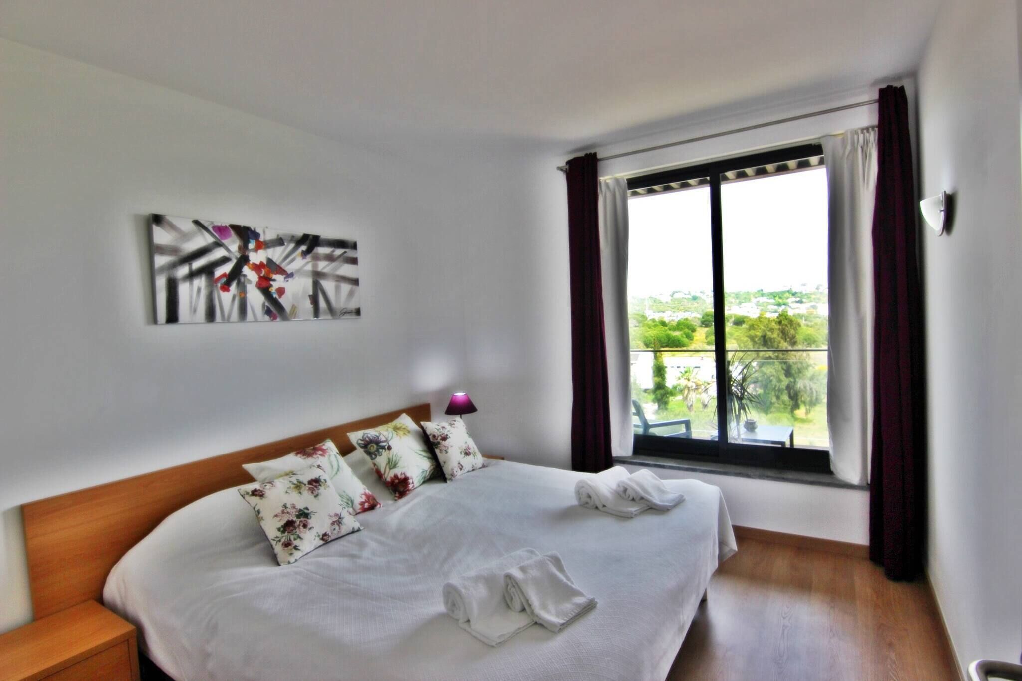 A hotel room with a bed, pillows, a painting on the wall, and a window with curtains.