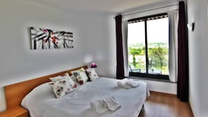 A hotel room with a bed, pillows, a painting on the wall, and a window with curtains. - Residences Golf Club - Pool and Tenis Court (Vilamoura)