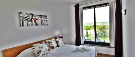 A hotel room with a bed, pillows, a painting on the wall, and a window with curtains.