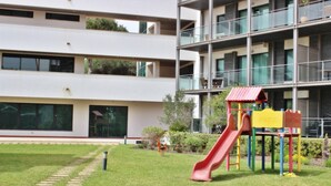A playground with a red slide and a yellow play structure, surrounded by a well-maintained lawn and a building with multiple floors and large windows. - Residences Golf Club - Pool and Tenis Court (Vilamoura)