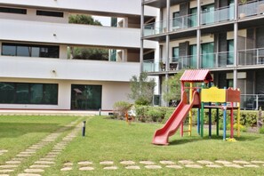 A playground with a red slide and a yellow play structure, surrounded by a well-maintained lawn and a building with multiple floors and large windows. - Residences Golf Club - Pool and Tenis Court (Vilamoura)