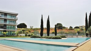 A swimming pool with a surrounding garden, a modern building with balconies, and a hilly landscape in the background. - Residences Golf Club - Pool and Tenis Court (Vilamoura)