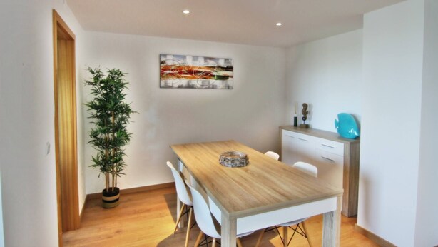 A modern dining area with a wooden table, white chairs, a potted plant, and a wall-mounted painting. - Residences Golf Club - Pool and Tenis Court (Vilamoura)