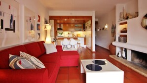 A living room with a red sofa, a white coffee table, and a fireplace. There is a kitchen in the background with wooden cabinets and a dining area. - Solar do Golfe - Pools & Old Village (Vilamoura)