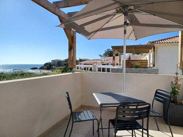 A balcony with a table and chairs, an umbrella, and a view of the sea and buildings.