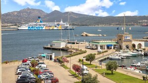 Un ferry llamado MOBY atracado en una marina con zona de estacionamiento, embarcaciones y un telón de fondo montañoso.