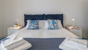Una cama bien tendida con sábanas blancas y dos almohadones decorativos, flanqueada por mesitas de noche blancas con lámparas y una pequeña planta.