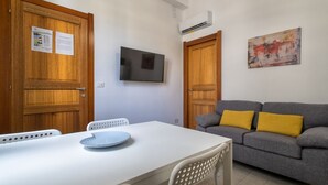 Ein Zimmer mit einem weißen Tisch, zwei Stühlen, einem grauen Sofa mit gelben Kissen, einem Wand-Fernseher und Holztüren.