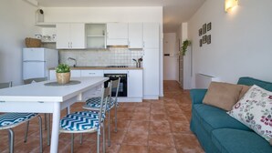 Een compacte keuken met witte kastjes, een eettafel met stoelen, en een marineblauwe bank met een bloemenkussen.