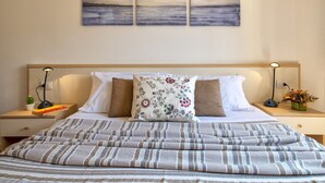 Een net opgemaakt bed met streepjesbeddengoed, een bloemenkussen en uitzicht op zee door drie schilderijen aan de muur.