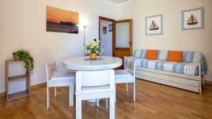 Une pièce avec une table de salle à manger ronde, des chaises blanches, un canapé avec des coussins à rayures, une petite plante sur une étagère, un tableau encadré représentant un coucher de soleil, et deux tableaux encadrés représentant des voiliers accrochés au mur.