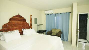 A hotel room with a large bed, a wooden headboard, a small table, a chair, and a door leading to another room.