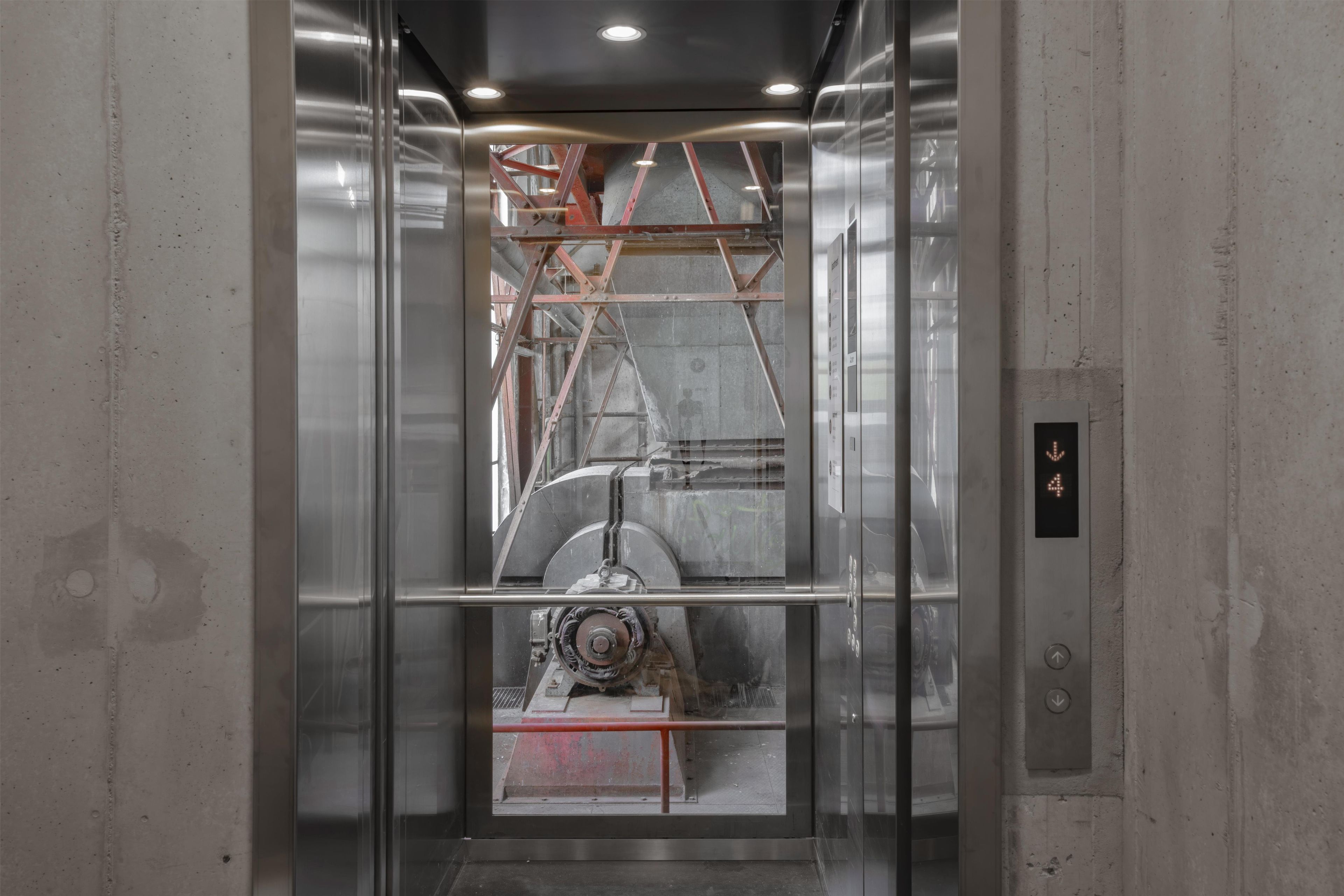 L’intérieur d’un ascenseur avec des parois métalliques, une porte en verre et un élément mécanique visible.