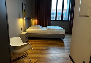 A modern bedroom with a bed, a white chair, a bedside table, and a window with curtains. - ipartment Leverkusen @HOME by Best Western (Leverkusen)
