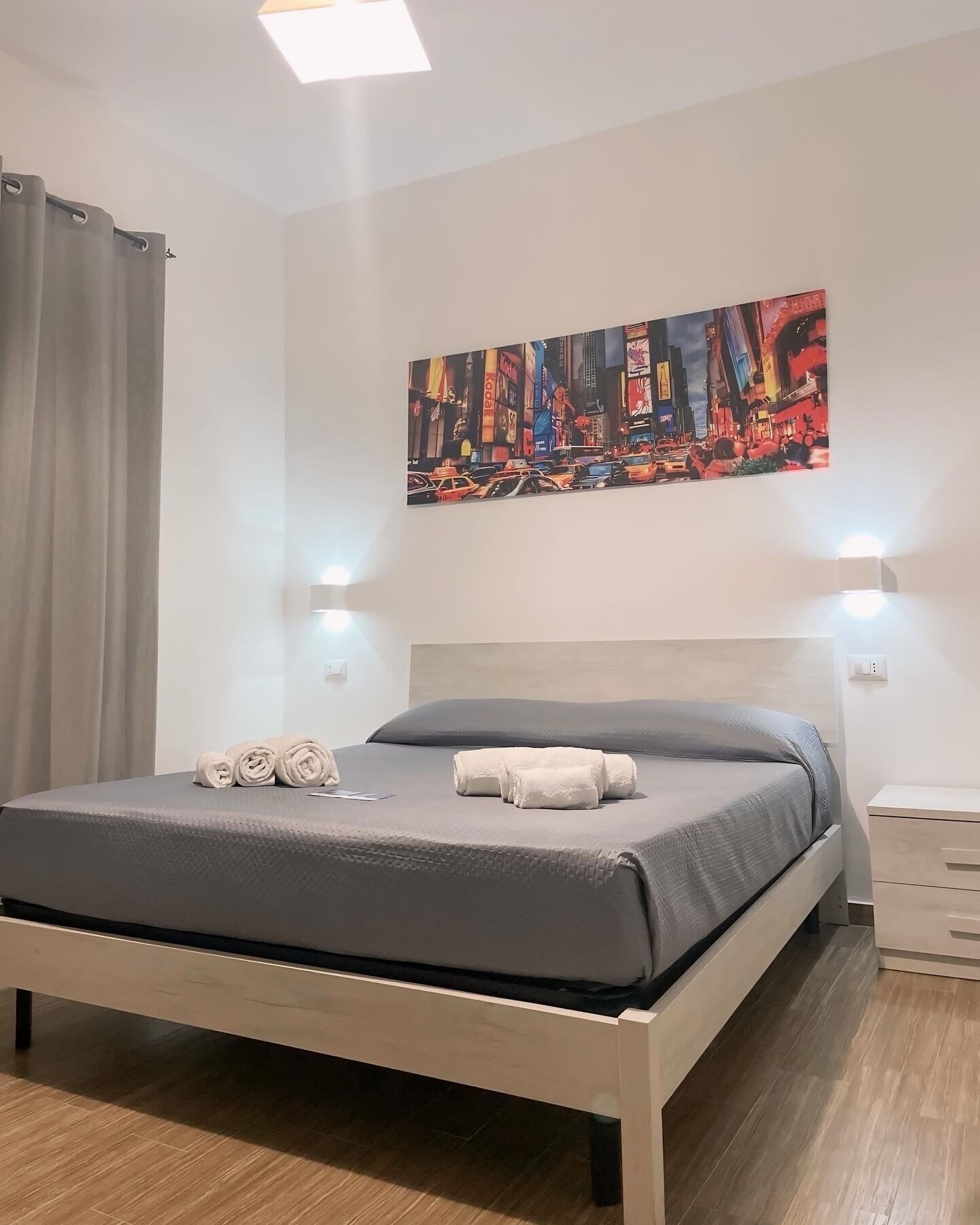 Una cama bien hecha con colchón y almohadas grises, un cuadro de un paisaje urbano en la pared y suelo de madera.