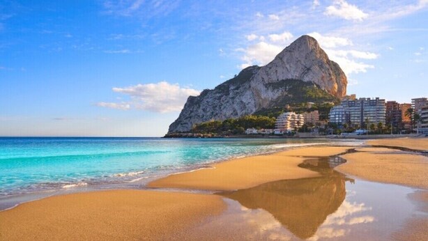 A beach with a large rock formation, clear blue water, and buildings in the background. - Home SEA&DREAMS Beach (Calp)