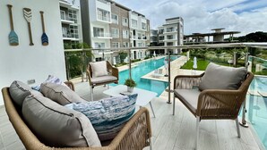 A balcony with wicker furniture, a pool, and a view of modern buildings. - Coral Bay by DR Collection (Punta Cana)