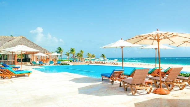 A poolside area with lounge chairs and umbrellas, a clear view of the ocean, and a thatched-roof structure. - Coral Bay by DR Collection (Punta Cana)
