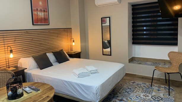 A hotel room with a bed, bedside lamps, a mirror, a window with blinds, a wicker chair, and a patterned rug. - Home SEA&DREAMS OldTown IV (Calp)