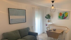 Una sala de estar con un sofá verde, una mesa de comedor de madera con sillas, una planta en maceta y un cuadro abstracto colorido.