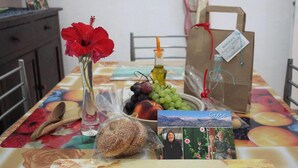 A table with a variety of food items, a vase with a red flower, a bottle of olive oil, and a gift bag with a tag.