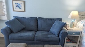 A blue sofa with three grey ottomans in front, a white bedside table with a lamp, and two framed pictures on the wall.